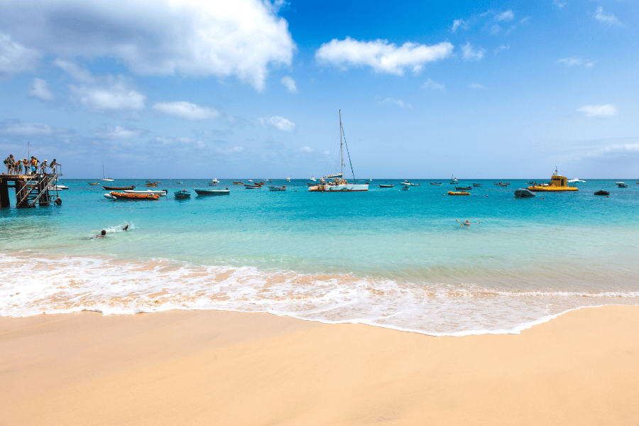 O pontão de madeira da Praia de Santa Maria rodeado por barcos de pesca locais e banhistas nas águas calmas e límpidas da ilha do Sal.